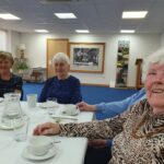 Three smiling people with grey hair sit around a table. The table is covered by a pale tablecloth with white crockery, including cups of tea on it