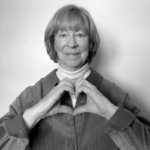 A greyscale image of a smiling person with short light coloured hair and wearing a light-coloured turtle neck and a dark coloured shirt over it. They are forming a heart symbol with their two hands
