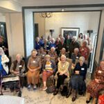 A group of smiling people sitting in beige high-backed chairs and others standing behind them. They are wearing smart casual clothing in autumn tones, and each has a white sticker on their chest. The room they are in has cream walls, black wooden doorways and a chandelier.