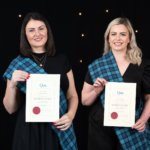 A colour image of two smiling people. One has long black hair, the other long blonde hair. They both wear a tartan sash, and each holds a white certificate with a red wax circle at bottom left