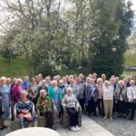 A large group of older adults gathered outdoors, standing and seated in front of blooming trees and greenery. Many are smiling at the camera, dressed in a variety of colourful and patterned clothing. The setting is a paved area with wicker chairs under a bright, partly cloudy sky.
