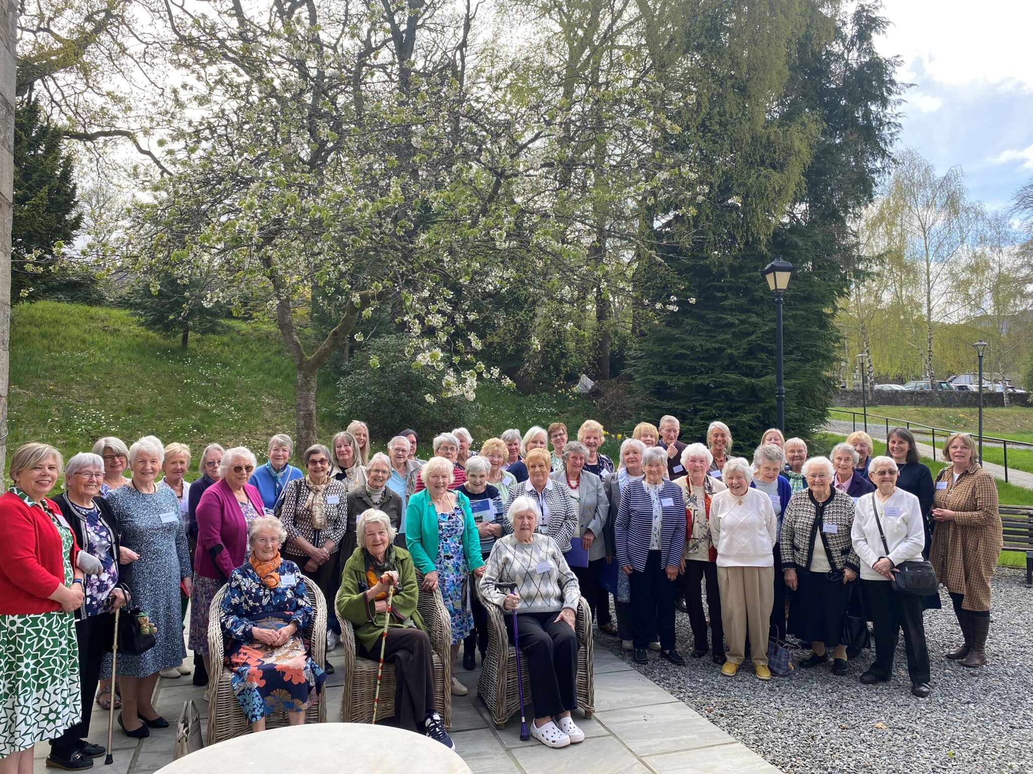A large group of older adults gathered outdoors, standing and seated in front of blooming trees and greenery. Many are smiling at the camera, dressed in a variety of colourful and patterned clothing. The setting is a paved area with wicker chairs under a bright, partly cloudy sky.
