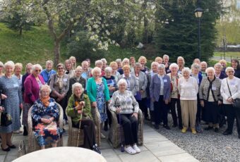 A large group of older adults gathered outdoors, standing and seated in front of blooming trees and greenery. Many are smiling at the camera, dressed in a variety of colourful and patterned clothing. The setting is a paved area with wicker chairs under a bright, partly cloudy sky.