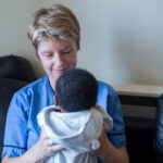 A person with short dark blonde hair wearing a blue nurse's uniform sits on a black leather sofa. They are smiling at a black haired baby they are holding