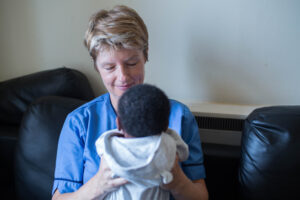 A person with short dark blonde hair wearing a blue nurse's uniform sits on a black leather sofa. They are smiling at a black haired baby they are holding