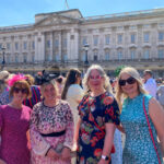 Four people dressed in vibrant, formal summer attire stand smiling in front of Buckingham Palace on a clear, sunny day. Each person is wearing a unique outfit with bright patterns and floral designs, along with decorative fascinators or headpieces. Two - far left and on the right - wear sunglasses and all appear to be enjoying the festive atmosphere, surrounded by a well-dressed crowd gathered outside the ornate palace gates.