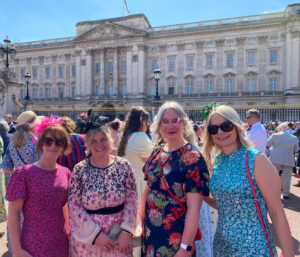 The image shows four people standing in front of Buckingham Palace. They are all smiling.