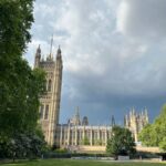 A colour image of the view of the Palace of Westminster in London, showing the Victoria Tower on the left and the ornate Gothic architecture of the Houses of Parliament. The building is framed by green trees on both sides, with a grassy park area in the foreground and a dramatic sky with dark clouds above.