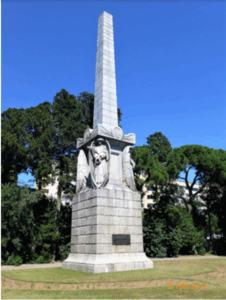 A large white stone tower-like monument on grass with trees and blue sky in the background